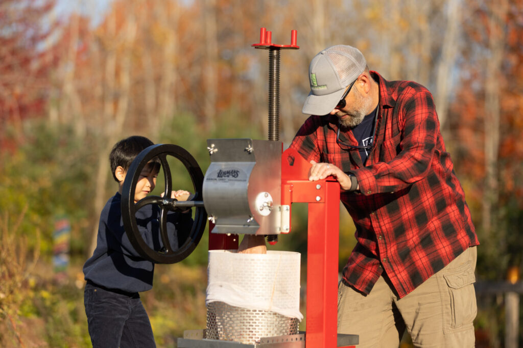 Young boy presses apples with adult mentor in autumn setting, capturing joy of learning.