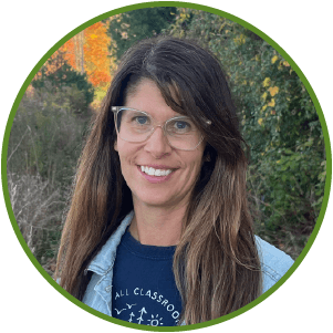 Smiling woman in glasses with cascading hair, dressed casually outdoors among colorful fall foliage.