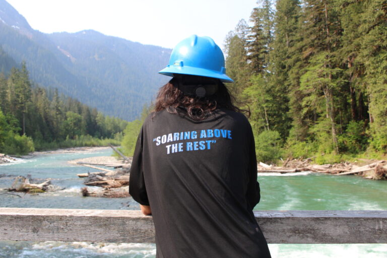 Person in hard hat gazes at turquoise river and mountains, inspiring exploration in nature.